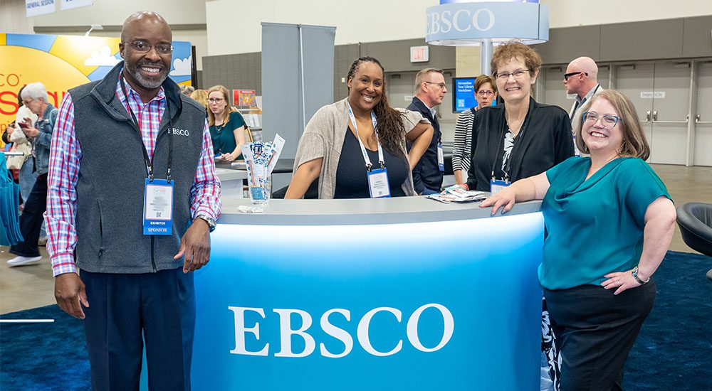 Group of one man and three women standing in front of sponsor EBSCO's booth.