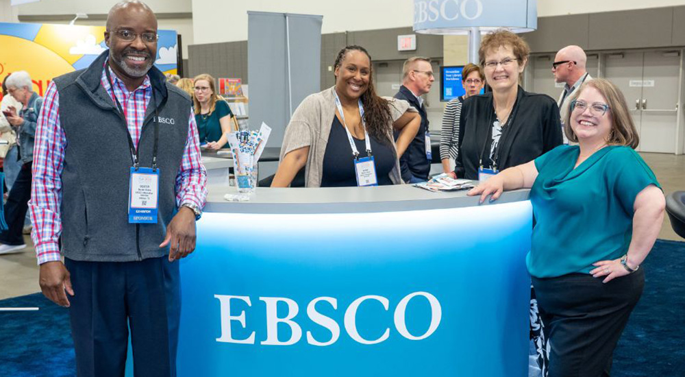Group of one man and three women standing in front of sponsor EBSCO's booth.