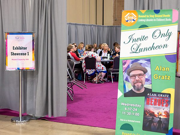 Outside of an Exhibitor Showcase with signs about the author and people seated at a table.
