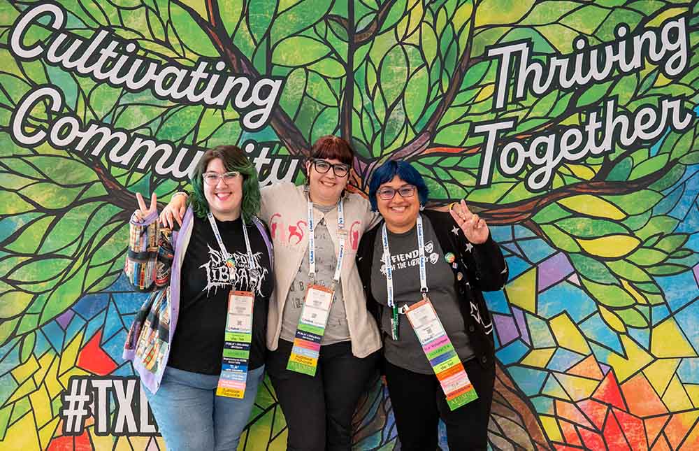 Three women in front to the TLA 2026 Selfie Wall of a mosaic tree with the words Cultivating Community Thriving Together.