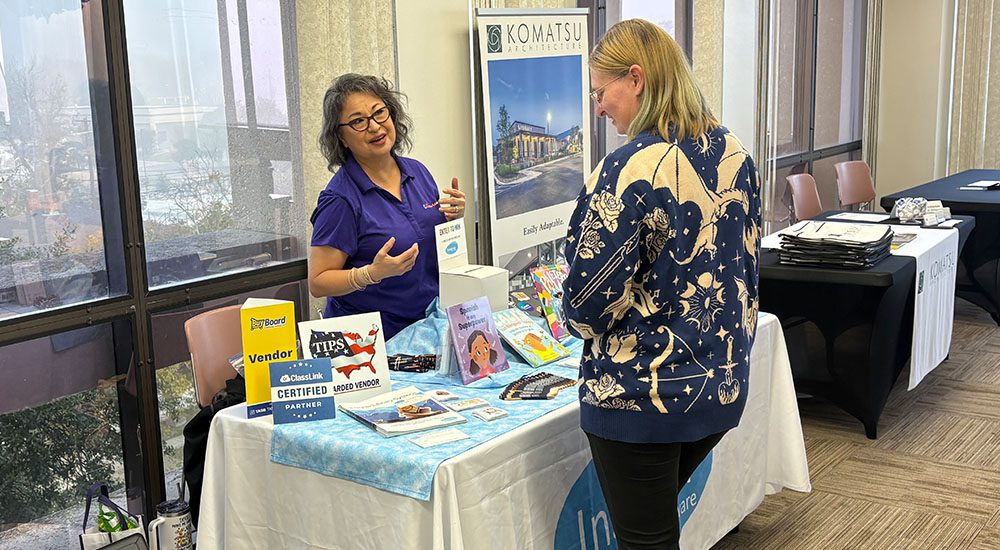 Two women at a conference, one behind a table and talking with an attendee